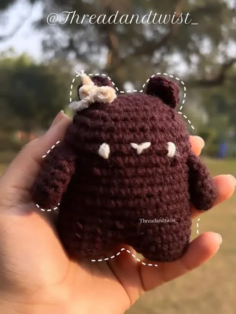 Small dark brown crochet bear with a flower, held in a palm